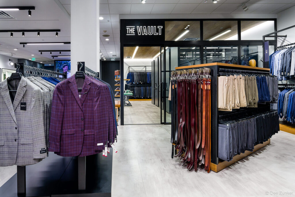 Interior view of The Vault menswear boutique showing suit jackets, belts, and trousers displayed on modern retail fixtures in Houston.