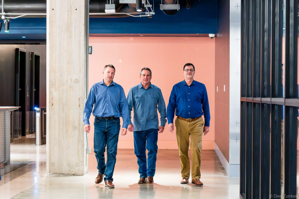 Three male executives in blue shirts walk confidently through a modern Houston office corridor, corporate branding photography by Dee Zunker.
