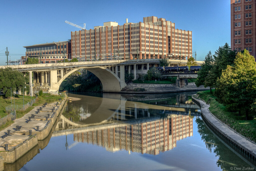 University of Houston brick building reflected in calm Buffalo Bayou waters beneath a concrete arch bridge, Houston Texas