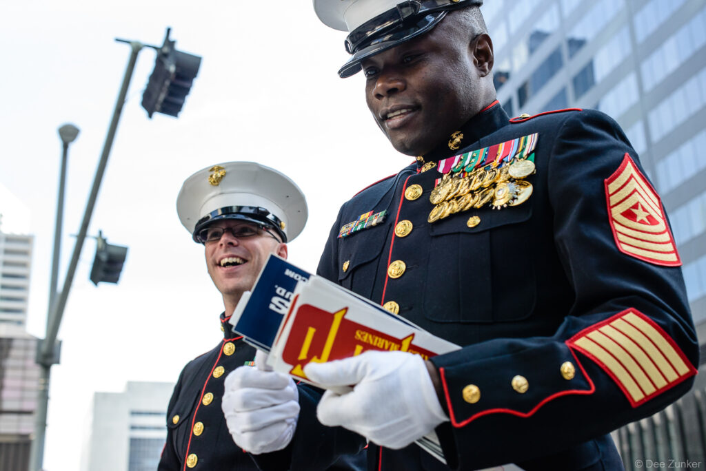 Two US Marines in dress blue uniforms with medals and white gloves at a Houston corporate event, holding printed materials.