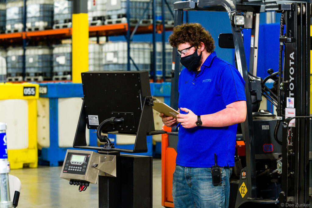 Warehouse worker in blue polo, safety glasses, and mask reviews clipboard at industrial kiosk beside a forklift in a Houston distribution facility.