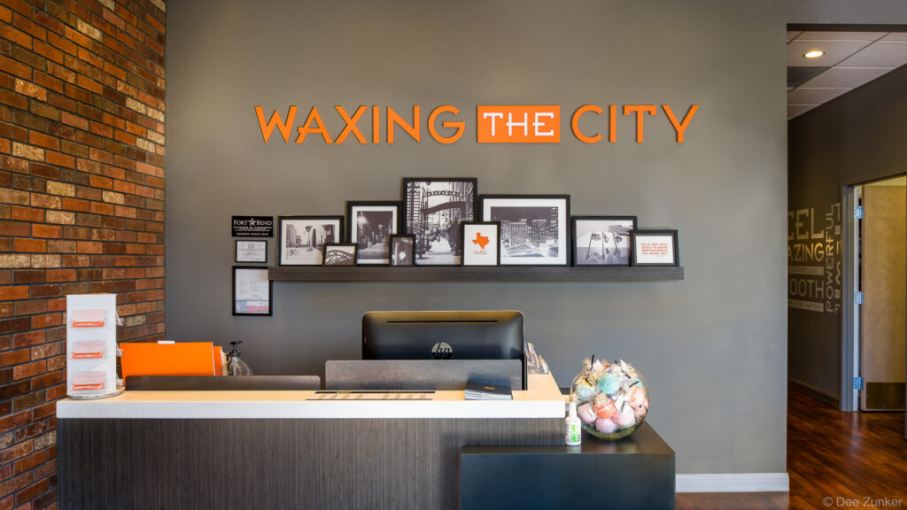 Modern hair removal studio reception desk with orange branded signage, framed city photos, and brick accent wall in Sugar Land TX