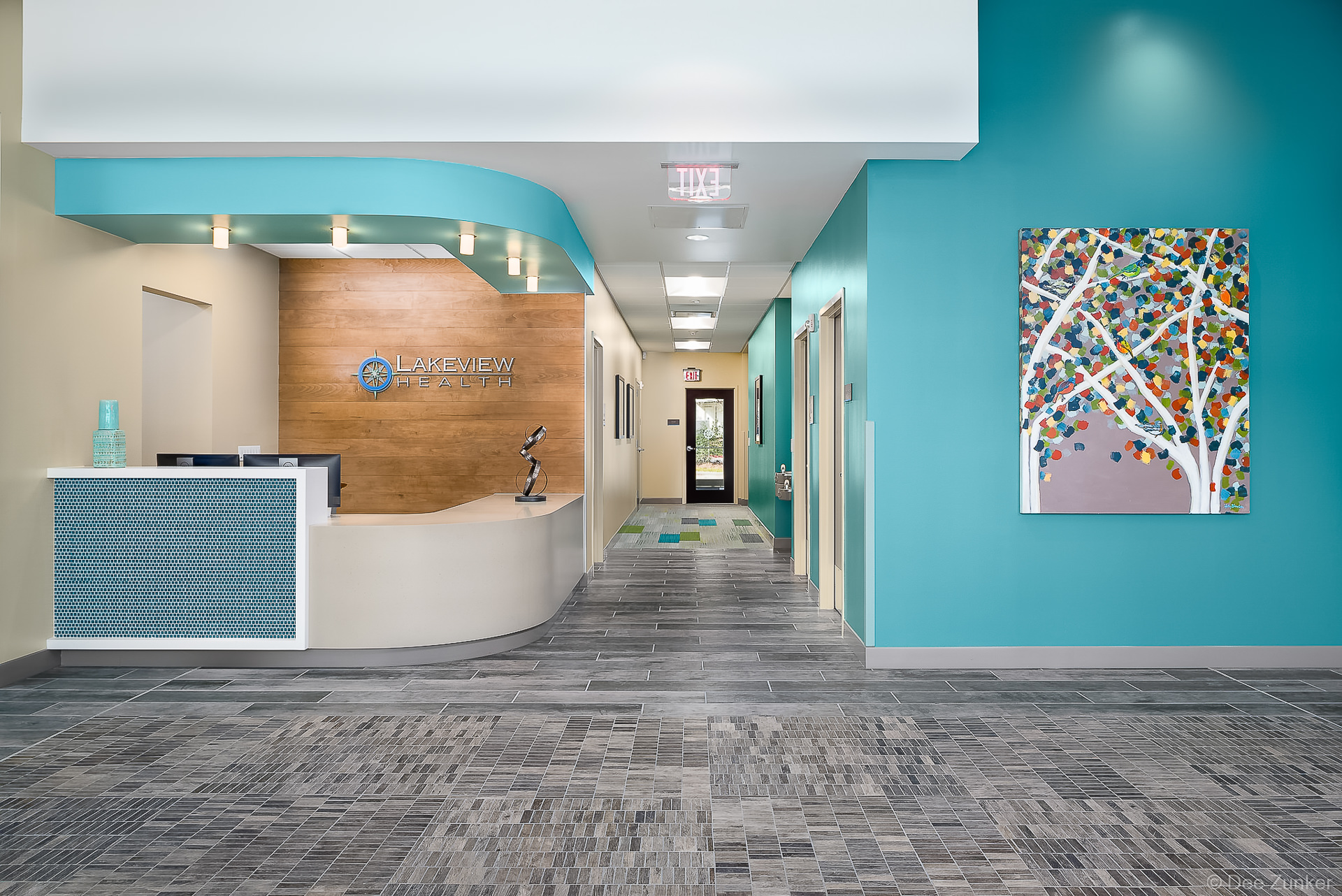 Modern healthcare facility reception area with teal accents, curved white desk, wood feature wall, and colorful tree artwork in The Woodlands TX